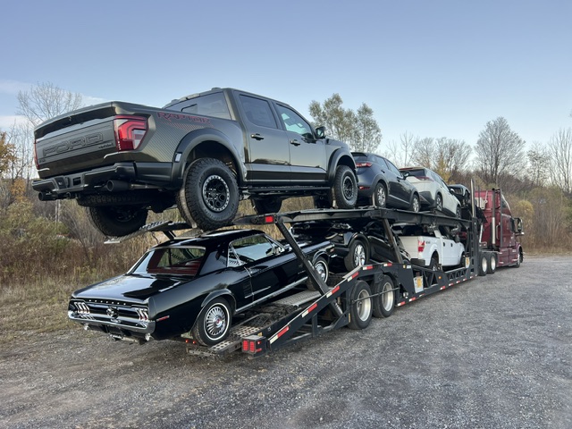 Maroon Allstar Hauling Volvo carrier loaded with a Ford F-150 Raptor, a classic black Mustang coupe, SUVs and a pickup on a gravel lot