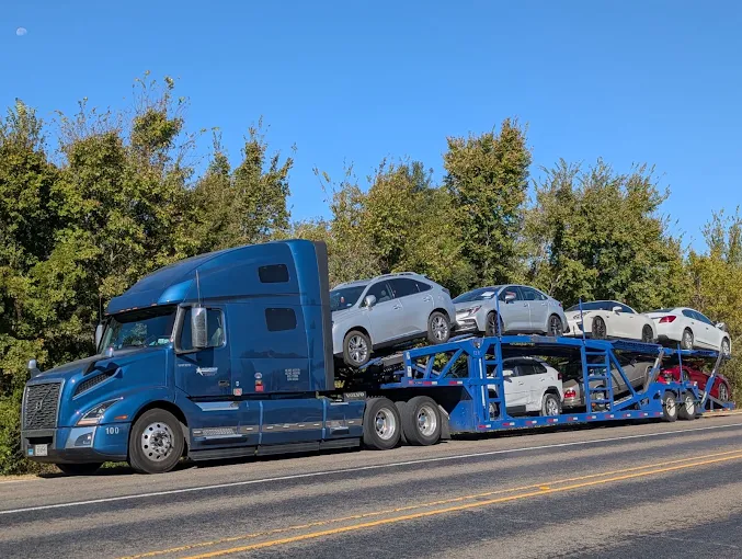 Blue Allstar Hauling Volvo VNL car hauler loaded with sedans and SUVs on a tree-lined highway
