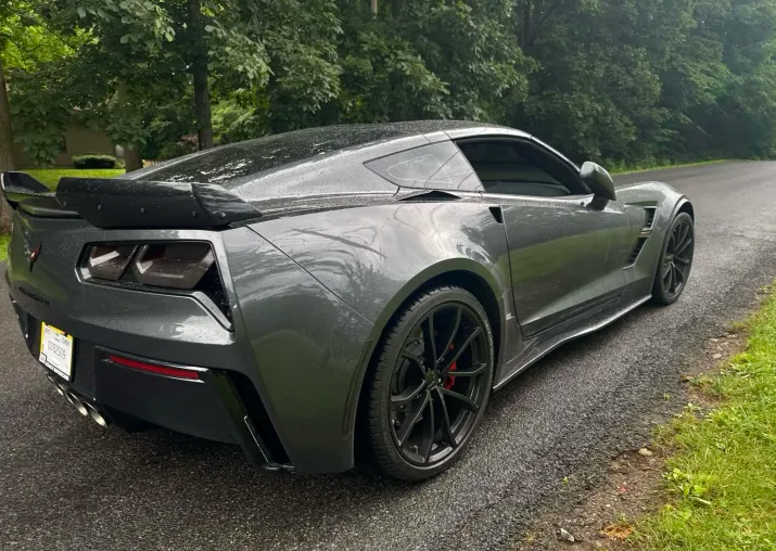 Dark gray Chevrolet Corvette C7 Z06 coupe with black multi-spoke wheels, red brake calipers and a carbon fiber rear wing parked on a wet roadside before Allstar Hauling transport
