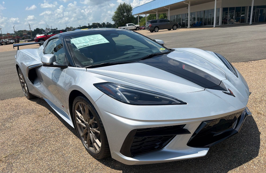 Silver Chevrolet Corvette C8 Stingray R package coupe with black roof and bronze wheels on a dealership lot before Allstar Hauling transport