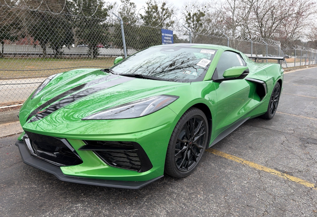 Bright green Chevrolet Corvette C8 Stingray coupe with black racing stripes and a high rear wing at an auction lot before Allstar Hauling transport