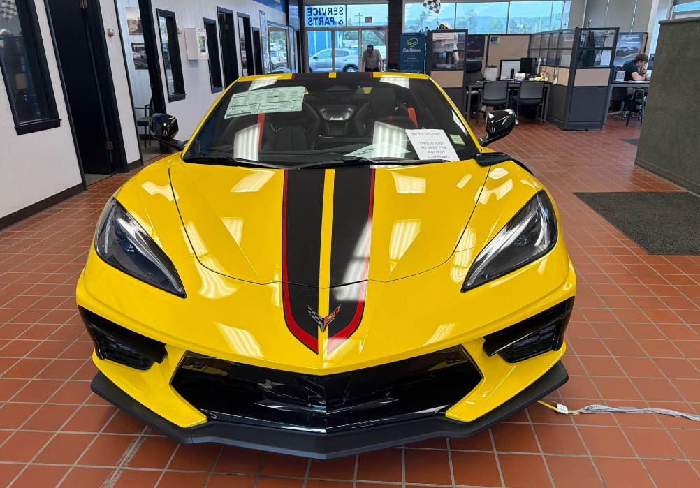 Yellow Chevrolet Corvette C8 Stingray coupe with black and red racing stripes inside a dealership showroom before Allstar Hauling transport