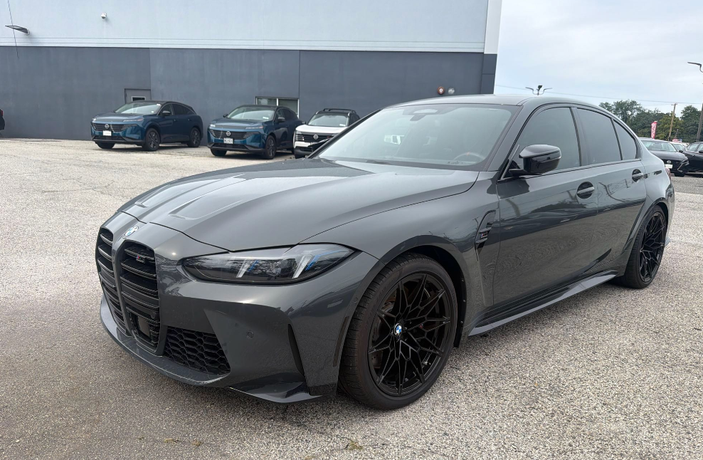 Charcoal gray BMW M3 sedan with black wheels staged for transport on a dealership lot